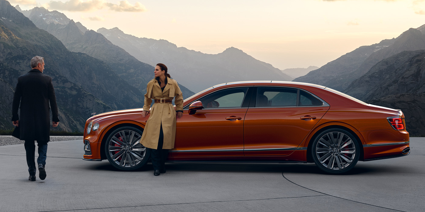 Bentley Polanco Bentley Flying Spur Speed parked in Orange Flame coloured exterior parked, with mountainous background and two people in view.